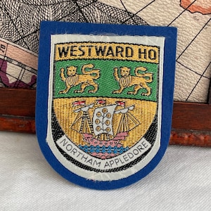 May include: A blue patch with embroidered text and imagery. The patch reads "WESTWARD HO" at the top and "NORTHAM APPLEDORE" at the bottom. The center features two golden lions above a ship.