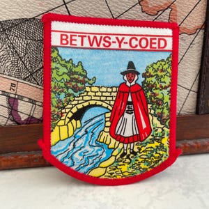 May include: A rectangular patch with a red border and the text "BETWS-Y-COED" at the top. The patch features a colourful illustration of a person in a red cloak standing near a bridge over a river.