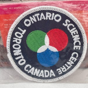 Könnte beinhalten: Gestickter Aufnäher mit dem Text "TORONTO ONTARIO SCIENCE CENTRE CANADA" in Weiß auf marineblauem Hintergrund. Der Aufnäher zeigt drei sich überlappende Kreise in Grün, Rot und Blau mit weißem Rand.
