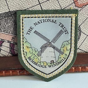 May include: A shield-shaped embroidered patch with a dark green felt border and yellow stitching. The centre features a light blue illustration of the Bembridge Windmill surrounded by trees, with the text THE NATIONAL TRUST BEMBRIDGE WINDMILL.