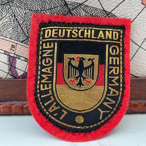 Puede incluir: Un parche rojo, negro y dorado con la palabra "DEUTSCHLAND" en la parte superior y "GERMANY" en el lado derecho. El parche presenta el escudo de armas alemán y las palabras "L'ALLEMAGNE" en el lado izquierdo.