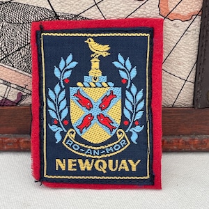 Op de afbeelding: Een rechthoekige patch met een marineblauwe achtergrond en een rode rand. De patch toont een gele vogel op een voetstuk, een schild met rode vissen en de tekst "NEWQUAY" in geel.