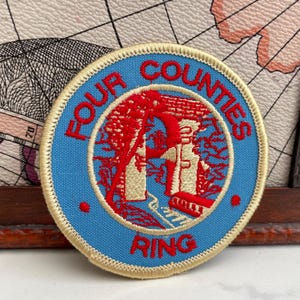 Puede incluir: Parche redondo con el texto "FOUR COUNTIES RING" en rojo y azul. El parche presenta una ilustración roja de un edificio y árboles en el centro, sobre un fondo azul claro y con un borde dorado.