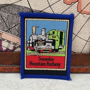 May include: A rectangular embroidered patch with a blue border featuring a steam locomotive in shades of gray, green, and black against a light blue sky. Below the train, a red banner contains the black text Snowdon Mountain Railway.