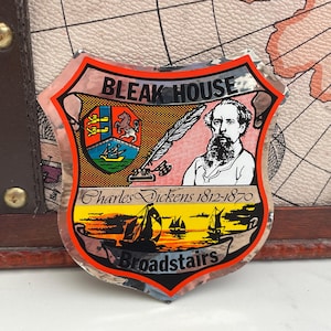 May include: A shield-shaped sticker with the words "Bleak House" at the top. The sticker features a portrait of Charles Dickens, a quill, and a coastal scene with sailboats. The text "Broadstairs" is at the bottom.