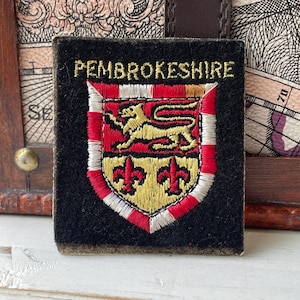 May include: A rectangular black fabric patch featuring the embroidered text PEMBROKESHIRE in gold above a shield. The shield displays a gold lion on a red background and two red fleur-de-lis on a gold background, bordered by red and white stitching.