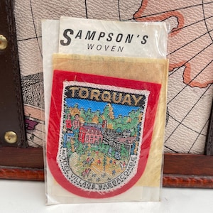 May include: A vintage Torquay patch with a red border, featuring a colorful illustration of a town scene. The patch is packaged with a paper label that reads "SAMPSON'S WOVEN". The patch is likely a souvenir or collectible item.