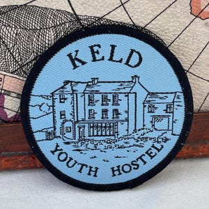 May include: A round patch with a light blue background and a black border. The patch features a detailed illustration of a building with the text "KELD YOUTH HOSTEL" in black. The design is reminiscent of a vintage travel souvenir.