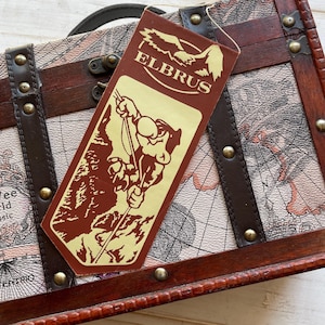 Könnte beinhalten: Ein brauner Lederkoffer mit einem Kartenmotiv und einem braunen Etikett mit der Aufschrift "ELBRUS" mit einer Cartoon-Abbildung einer Person, die einen Berg besteigt.