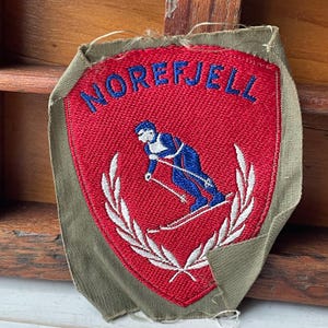 May include: Embroidered patch featuring the word "NOREFJELL" in blue on a red shield. It depicts a skier in blue and white, with a white laurel wreath. The patch is edged with olive green fabric.