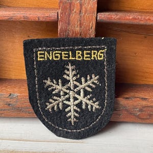 May include: Black felt patch with the word "ENGELBERG" in gold thread above a snowflake design. The patch has a stitched border and is set against a wooden background.