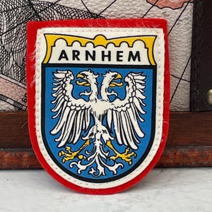 May include: A shield-shaped embroidered patch featuring a double-headed eagle in white with yellow talons on a blue background. The word ARNHEM is printed in black capital letters on a white banner at the top, all bordered by a red felt edge.