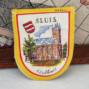May include: A shield-shaped patch with a yellow border and a white interior. The patch features the word "SLUIS" above a detailed illustration of a building with a red roof and a church tower. A red and white emblem is on the left, and the word "stadhuis" is at the bottom.