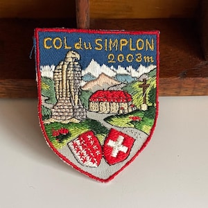 May include: Embroidered patch with a shield shape featuring the text "COL du SIMPLON 2003m". The patch depicts a stone structure, a red-roofed building, and two crests. The background includes mountains and a blue sky.