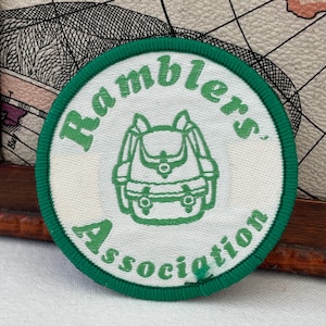 Op de afbeelding: Een ronde geborduurde patch met een witte achtergrond en een groene rand. Het midden bevat een groene lijntekening van een rugzak. De tekst "Ramblers' Association" is in het groen geborduurd en buigt om de rugzak heen.