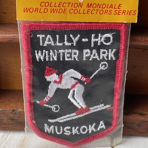 May include: A rectangular patch with a red border and the words "TALLY-HO WINTER PARK MUSKOKA" in white. The patch features an embroidered skier in white and red on a black background. The top of the patch reads "COLLECTION MONDIALE WORLD WIDE COLLECTORS SERIES".