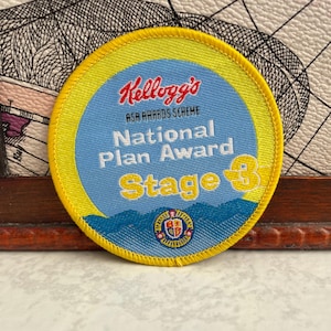 May include: A round, embroidered patch with a yellow border and a light blue center. The patch features the Kellogg's logo in red, the text "ASA AWARDS SCHEME", "National Plan Award", and "Stage 3". A small emblem is at the bottom.