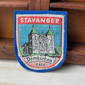 Könnte beinhalten: Ein blaues, gesticktes Abzeichen mit dem Wort "STAVANGER" oben. Das Abzeichen zeigt eine detaillierte Illustration einer Kirche mit grünbedachten Türmen und dem Text "Domkirken" und "12 ÅRH." in einem roten Rand.
