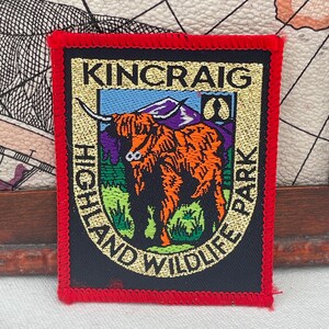 Może przedstawiać: Prostokątna naszywka z napisem "KINCRAIG HIGHLAND WILDLIFE PARK" w złotych literach. Naszywka przedstawia ilustrację szkockiej krowy w kolorze pomarańczowym i czarnym, z fioletową górą w tle. Naszywka ma czerwoną obwódkę.