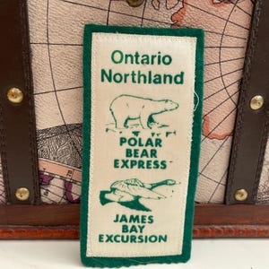 Puede incluir: Un parche rectangular crema con ribete verde presenta el texto "Ontario Northland" sobre una ilustración de un oso polar. Debajo, se lee "Polar Bear Express" y "James Bay Excursion". El parche es probablemente un recuerdo o artículo de viaje.