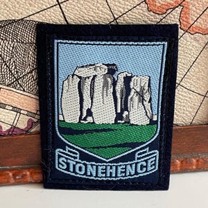 May include: A rectangular embroidered patch featuring a light blue shield shape with a dark blue border. The center depicts a detailed gray stone monument on a green field. Below the monument, a light blue banner contains the text STONEHENGE.