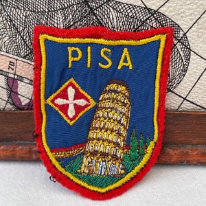 Könnte beinhalten: Ein schildförmiger, bestickter Aufnäher mit blauem Hintergrund und rotem Rand. Der Text PISA ist oben in Gelb eingestickt. Er zeigt den gelben Schiefen Turm von Pisa, ein rot-weißes rautenförmiges Emblem und grüne Bäume.