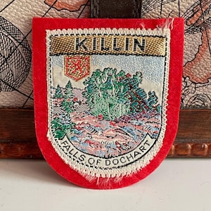May include: A shield-shaped fabric patch with a bright red felt border. The center features a woven landscape of the Falls of Dochart in Killin, Scotland, with a small red lion crest, green trees, and rocky terrain. The text reads KILLIN and FALLS OF DOCHART.