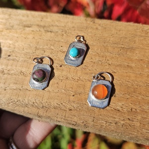 May include: Three silver charms with different colored gemstones. The charms are rectangular with a decorative border and a small loop at the top. The gemstones are turquoise, red, and orange.