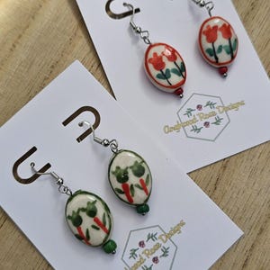 May include: Oval ceramic earrings with a floral design. The earrings have a white base with red and green painted flowers. The earrings are displayed on white cards with the text "Angharad Rose Designs".