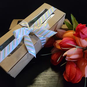 May include: A brown cardboard gift box with a window, tied with a pink and blue striped ribbon. The box is sitting on a dark wood surface next to a bouquet of pink and red tulips.