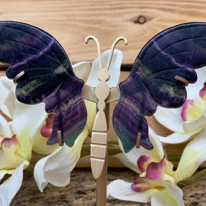 May include: A purple and green butterfly-shaped fluorite crystal on a gold stand. The crystal is polished and has a natural, organic look.