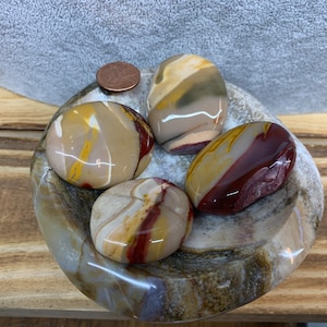 May include: Five smooth, polished stones with red, yellow, and brown swirling patterns. The stones are arranged in a bowl made of the same material.