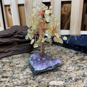 May include: A decorative tree sculpture with a copper wire trunk and branches, adorned with yellow gemstone leaves. The tree is mounted on a purple amethyst crystal base, creating a unique and natural aesthetic. The sculpture is a decorative item.