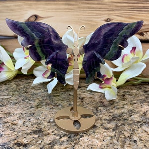 May include: A carved butterfly sculpture with purple and green wings is displayed on a gold-colored metal stand. The butterfly wings have a natural stone appearance. The stand has a circular base and a slender stem.