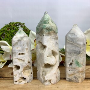 May include: Three white and green crystal points with a pointed top. The crystals have a rough, natural texture and are arranged in a row on a wooden surface.