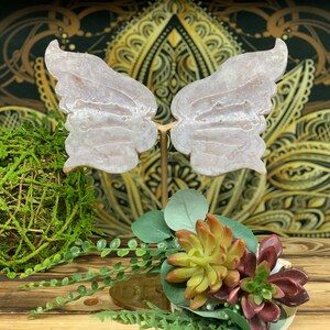 May include: A pair of white, translucent, butterfly-shaped wings made of polished stone, mounted on a gold metal stand. The wings are displayed against a background of gold and brown patterned fabric.