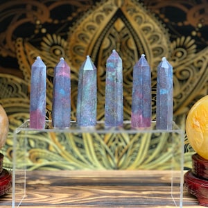 May include: Six iridescent purple and blue crystal wands on a clear display stand. The wands are approximately 2 inches long and have a faceted, pointed tip. The stand is on a wooden surface.