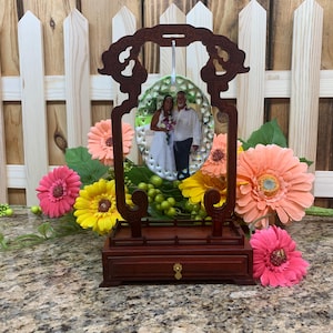 May include: A dark wood picture frame with an ornate top and a small drawer. The frame holds a photo of a couple. The frame is surrounded by colorful flowers, including pink, yellow, and orange gerbera daisies.