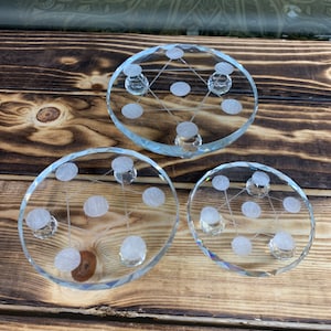 May include: Three clear glass crystal coasters with a geometric design and small crystal feet. The coasters are different sizes and are arranged in a row on a wooden surface.