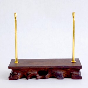 May include: A display stand featuring a rectangular dark brown wooden base with a natural, irregular root-like texture along the bottom edge. Two vertical, slender, polished gold-colored metal rods are attached to the top surface of the wooden base.