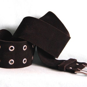 Peut inclure: Une ceinture en daim marron foncé avec un large bracelet à œillets argentés. La ceinture a une boucle argentée et est présentée sur fond blanc. Le bracelet est enroulé et la ceinture est drapée.