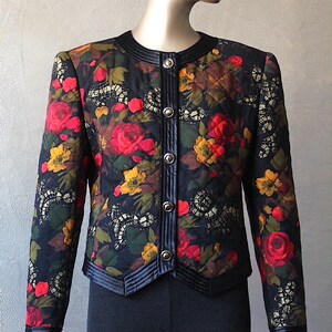 May include: A black quilted jacket with a floral print featuring red, yellow, and green flowers. The jacket has a black velvet collar and trim, and silver buttons down the front.