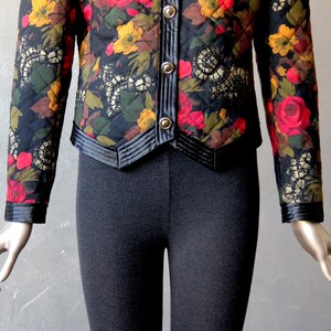 May include: A black quilted jacket with a floral print featuring red, yellow, and green flowers. The jacket has a black satin trim and a button closure.