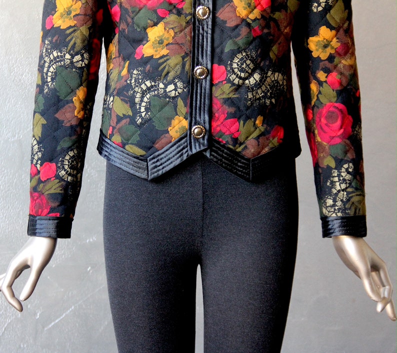 May include: A black quilted jacket with a floral print featuring red, yellow, and green flowers. The jacket has a black satin trim and a button closure.