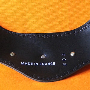 以下が含まれることがあります： 黒革製のベルトの裏側をクローズアップ。ベルトには「MADE IN FRANCE 304」と印刷されています。ベルトには銀色のリベットとステッチが施されています。