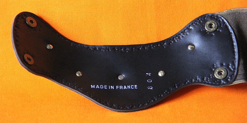 以下が含まれることがあります： 黒革製のベルトの裏側をクローズアップ。ベルトには「MADE IN FRANCE 304」と印刷されています。ベルトには銀色のリベットとステッチが施されています。