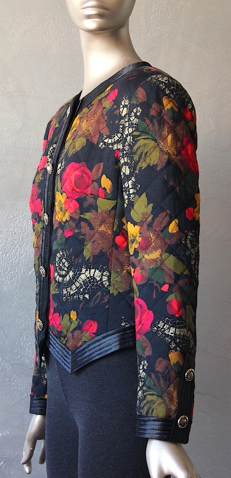 May include: A black quilted jacket with a floral print featuring red, yellow, and green flowers. The jacket has a black collar, black trim, and gold buttons.
