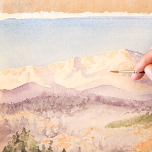 Op de afbeelding: Een hand houdt een penseel vast en voegt details toe aan een aquarel van een bergketen. De bergen zijn geschilderd in tinten paars, blauw en wit, met een lichtblauwe lucht op de achtergrond.