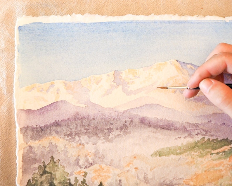 Op de afbeelding: Een hand houdt een penseel vast en voegt details toe aan een aquarel van een bergketen. De bergen zijn geschilderd in tinten paars, blauw en wit, met een lichtblauwe lucht op de achtergrond.
