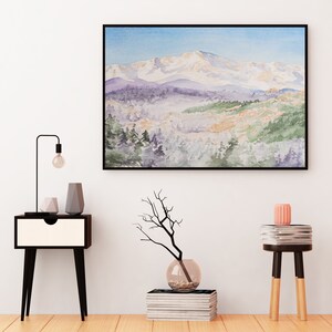 Op de afbeelding: Een ingelijst aquarel schilderij van een besneeuwde bergketen met een bos op de voorgrond. Het schilderij hangt aan een witte muur boven een zwart nachtkastje met een lamp en twee vazen. Een vaas met een tak en een stapel boeken staat op de vloer voor het nachtkastje. Een kleine tafel met een stapel boeken en een roze kaars staat aan de rechterkant van de afbeelding.